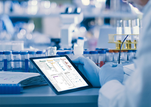 Photo of a hand where blue gloves and holding a tablet device in a laboratory setting, with blue-capped tubes in the background.